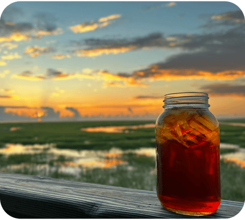 A mason jar filled with Savannah Sweet Tea set against a breathtaking backdrop of a marsh under a vibrant sunset, epitomizing the serene beauty of the South